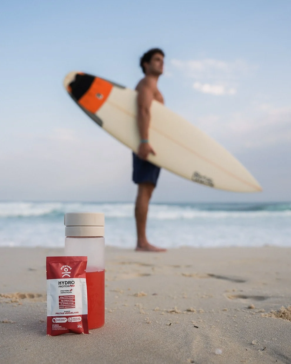 Hydro Protein PRO Frutas Vermelhas e garrafa na praia, com surfista ao fundo